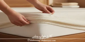 «نمای نزدیک دست‌ها هنگام کشیدن کاور روی تشک برای نمایش تفاوت کاور تشک و محافظ تشک در اتاق خوابی با توناژ کرم و نور گرم»